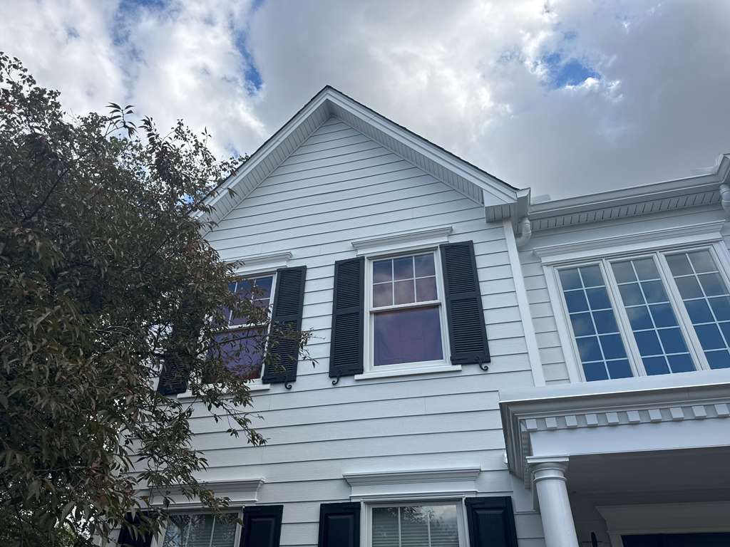 White two-story house exterior with light-colored horizontal siding, black window shutters, and a peaked roof under a sky.