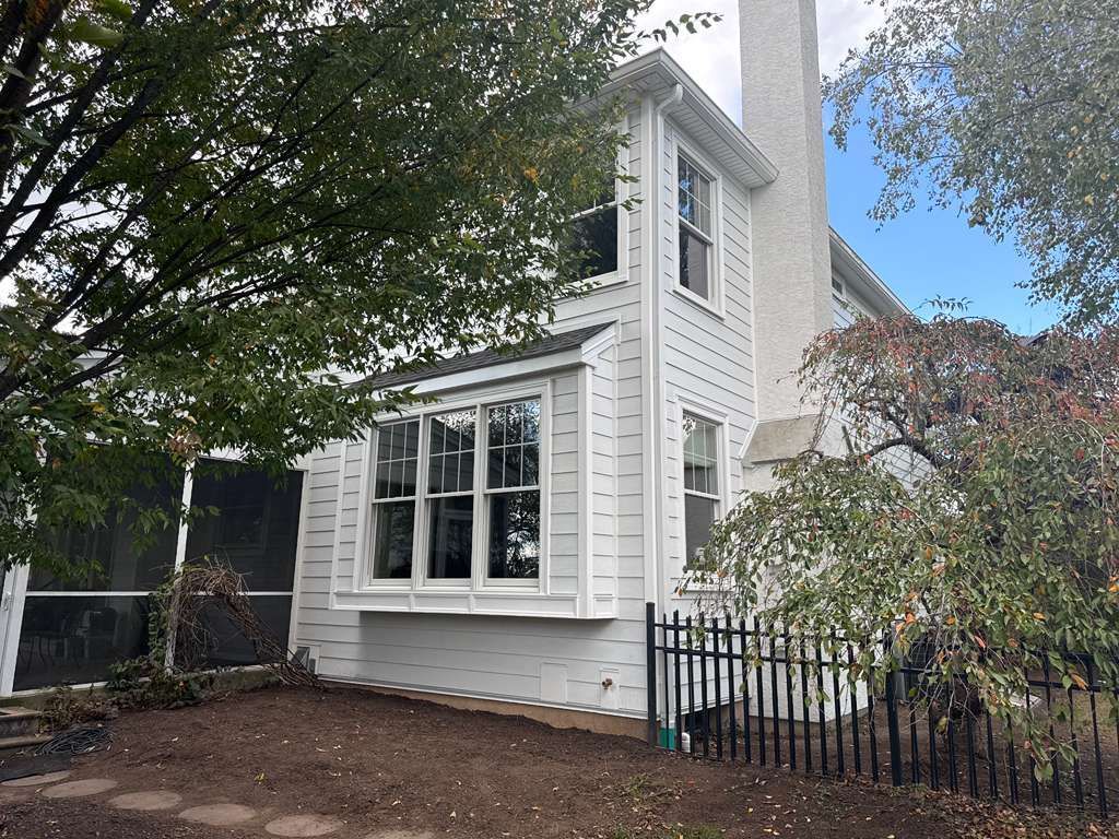 White two-story house with a bay window, chimney, and screened porch, framed by trees on a sunny day.