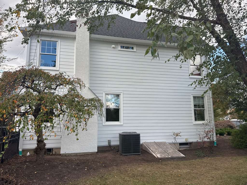 Two-story white house with a chimney, a tree in the foreground, an air conditioning unit, and a basement bulkhead door.