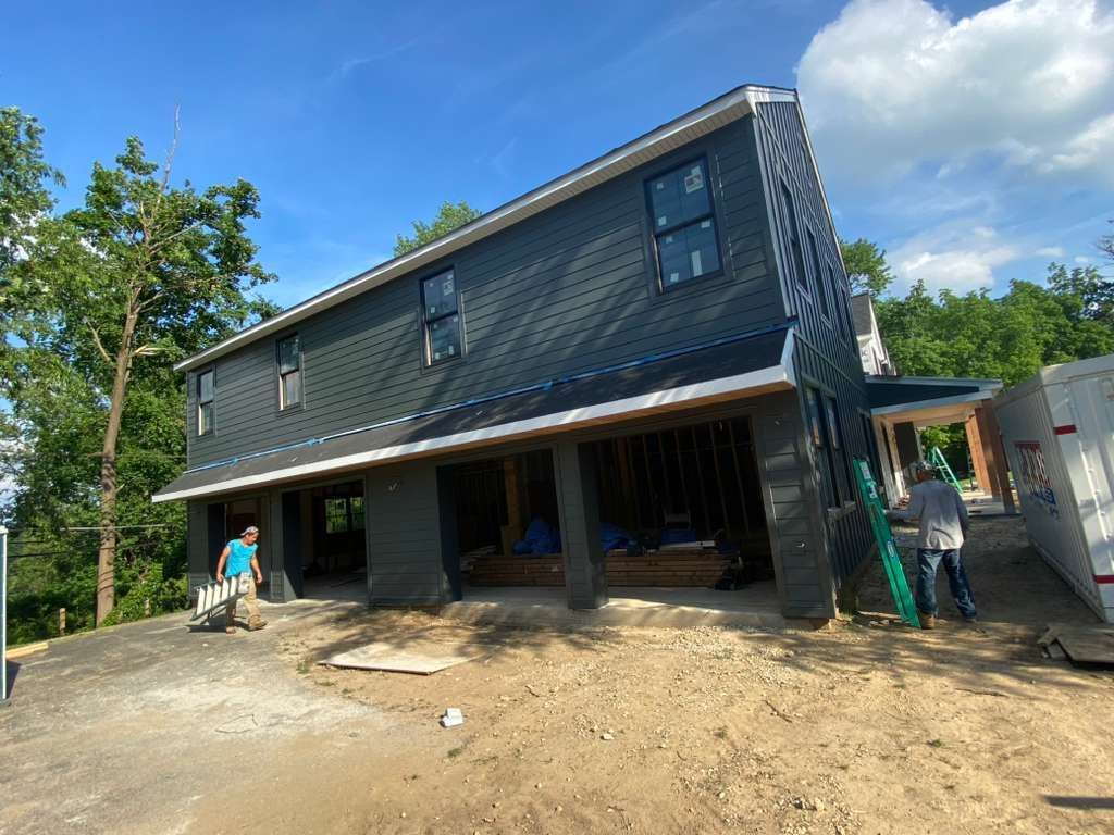 Two people work on the exterior construction of a dark-gray two-story house with an open ground-level garage space.