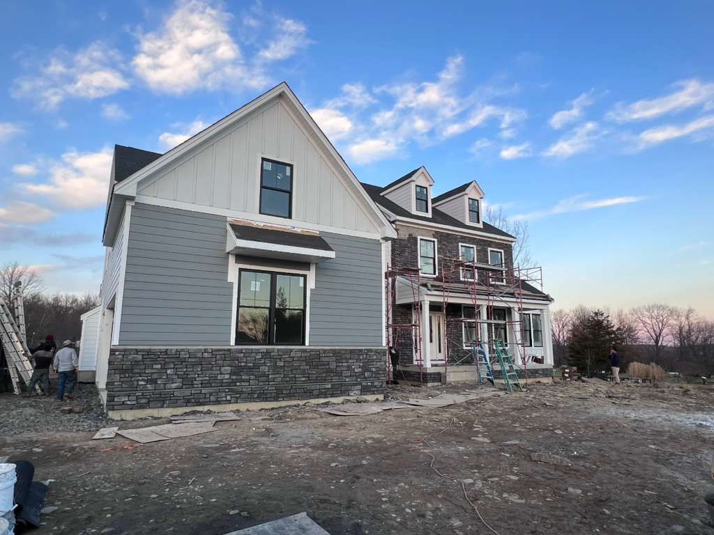 A two-story house under construction with grey horizontal siding, white vertical siding, and stone veneer at the base.