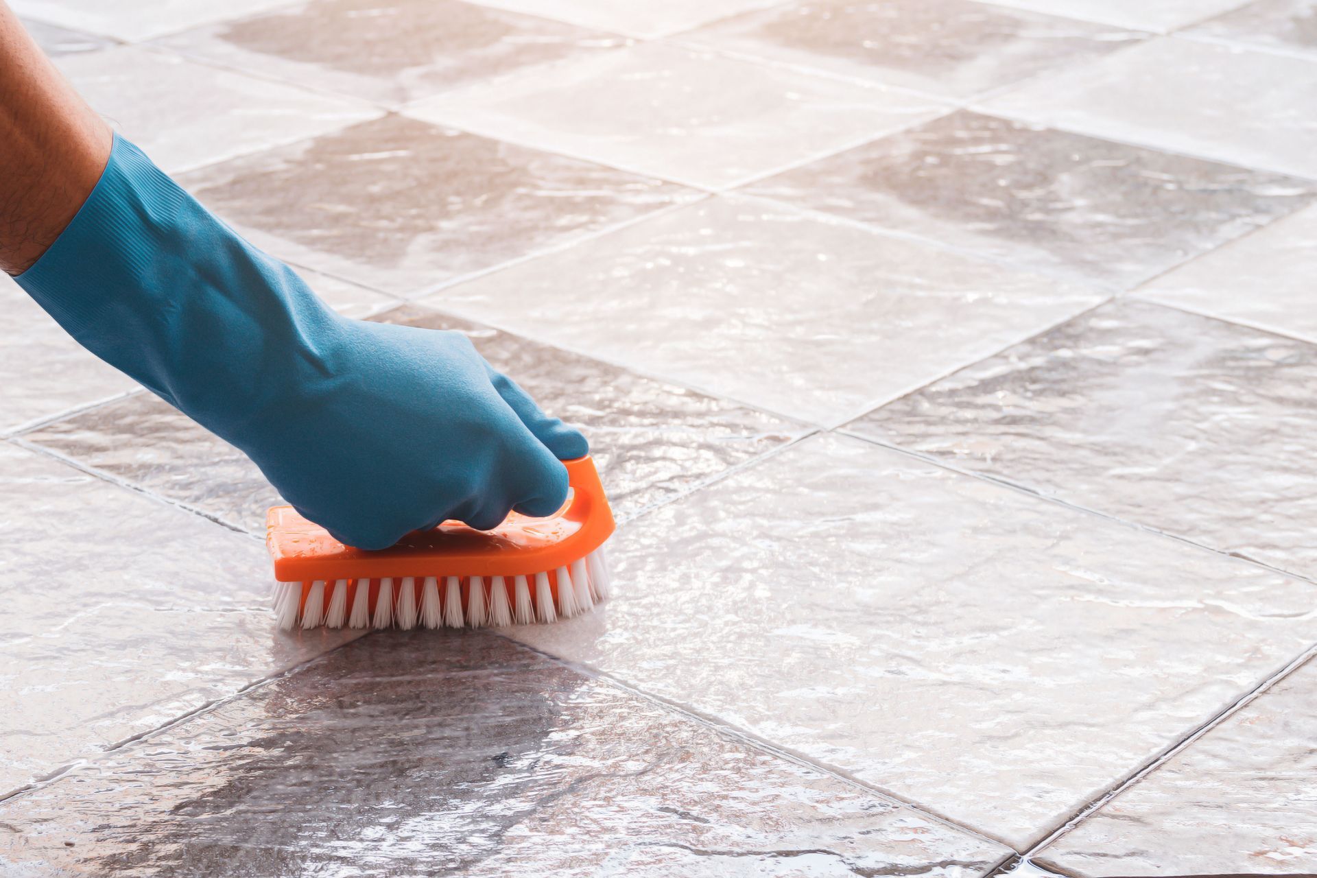 Hand in blue glove scrubbing tiled floor with orange brush.