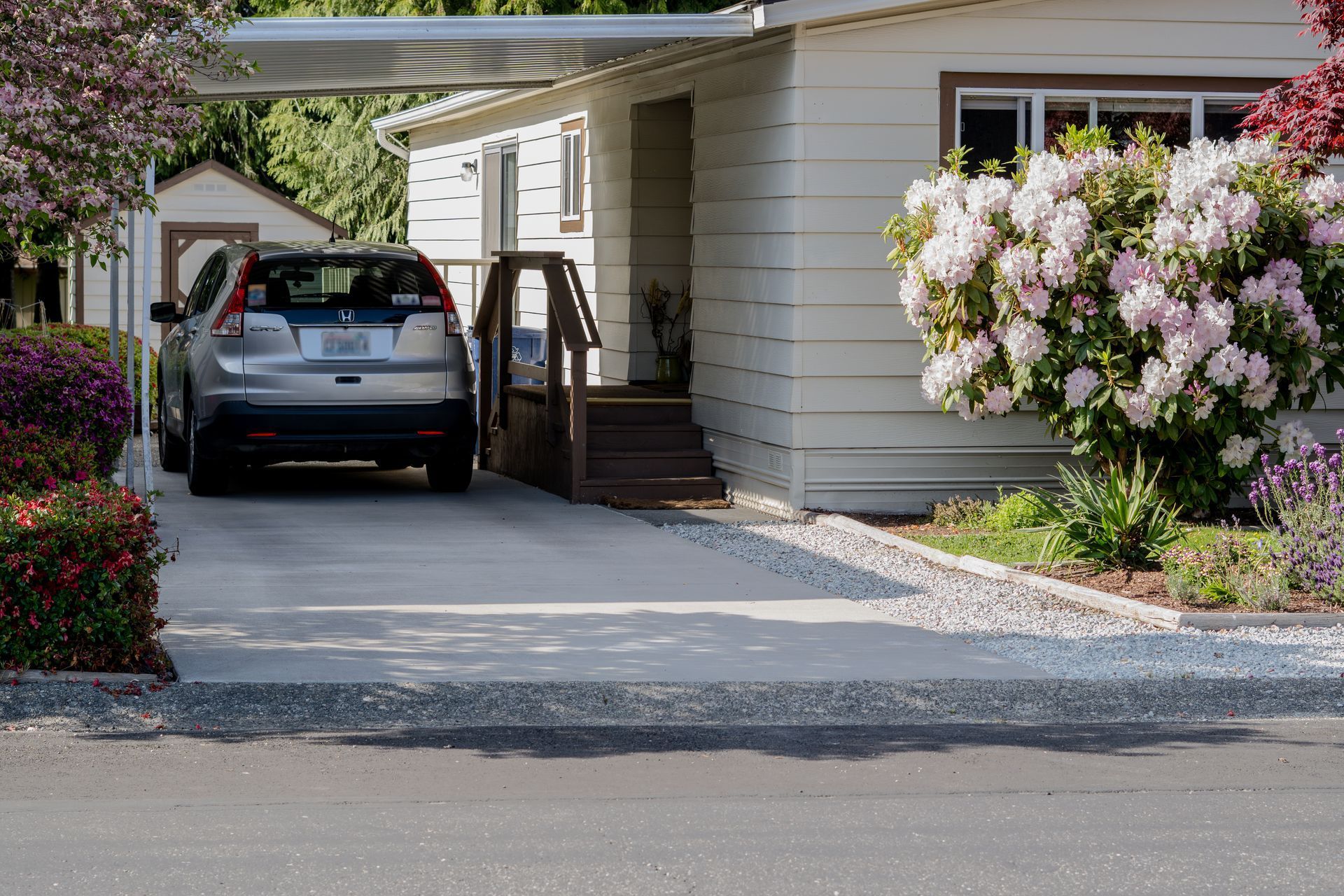 Carport with a parked SUV beside a house and flowering shrubs along the driveway
