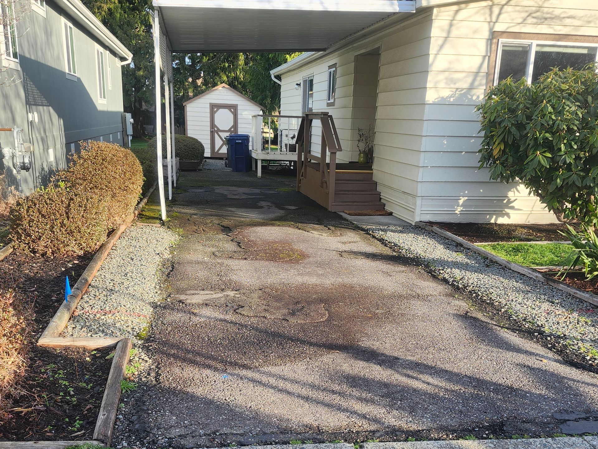 Narrow gravel driveway between houses, with bushes, a carport, and a small shed at the end.