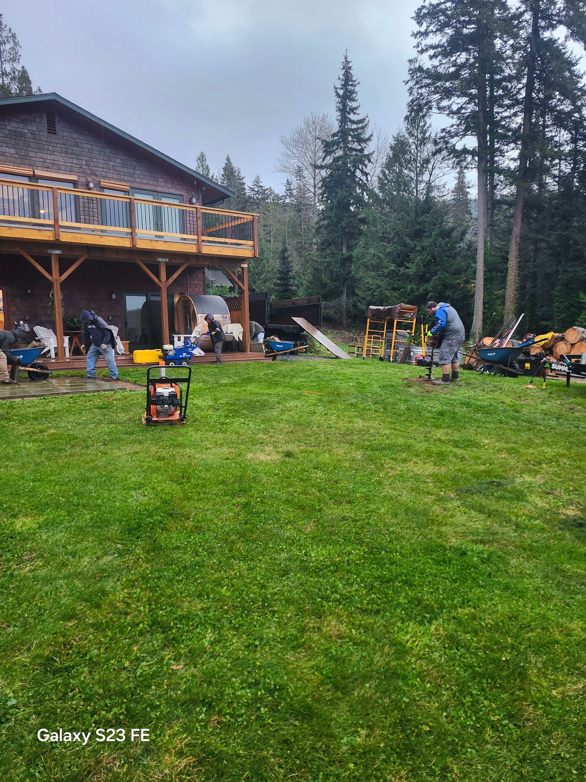 Backyard gathering beside a log cabin, with people, lawn chairs, and a grassy yard under overcast sky