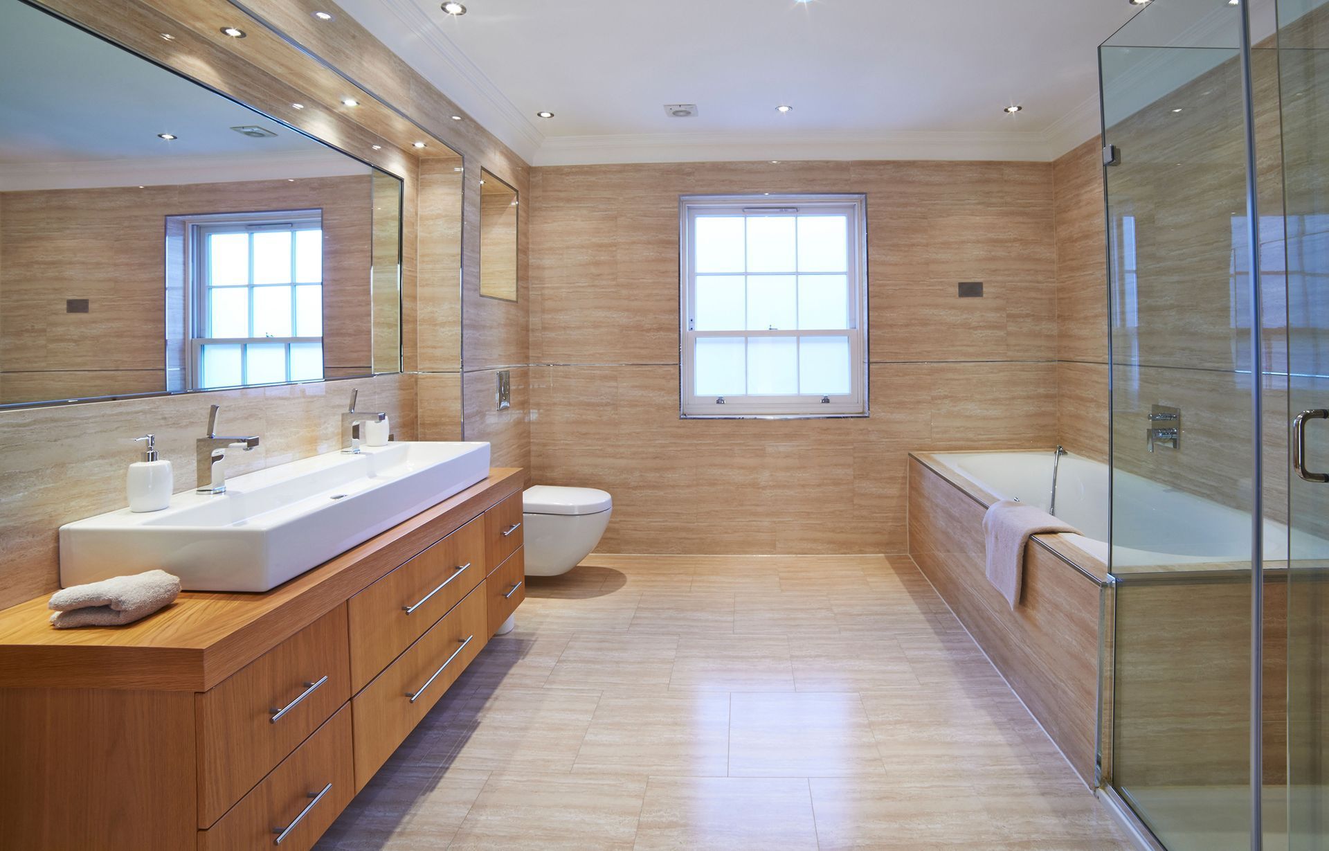 Bathroom with beige tile walls and floor, wood vanity, large mirror, white bathtub, and glass shower.