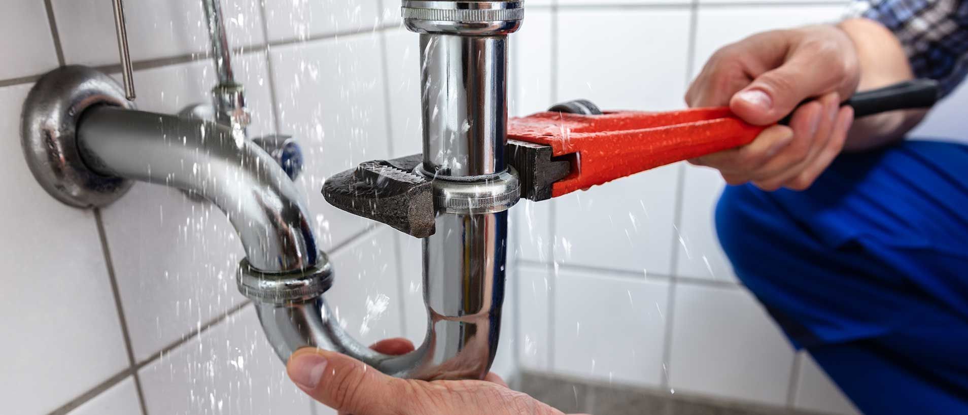 A plumber uses a wrench on a leaking pipe in a bathroom. Water sprays.