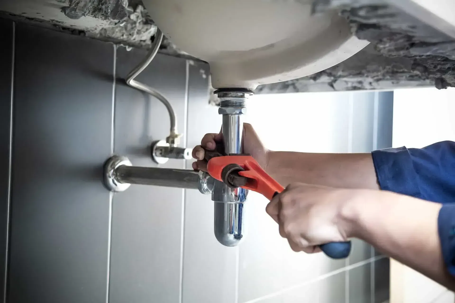 Plumber using a wrench to tighten a pipe under a bathroom sink; gray tiles on the wall.