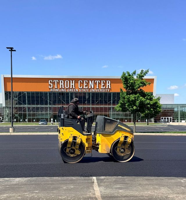 Yellow road roller parked in front of Stroh Center under a clear blue sky