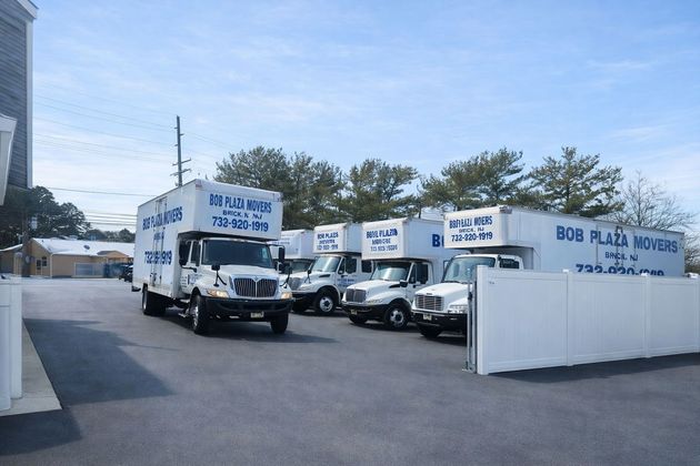 Moving trucks parked outside a building, advertising "Bob's Place Movers". Blue sky, white fence, and trees in the background.