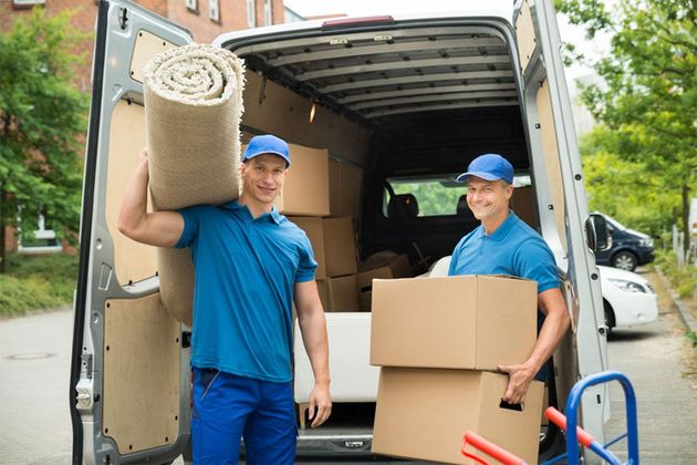 Two movers unloading boxes and a rolled carpet from a truck outdoors