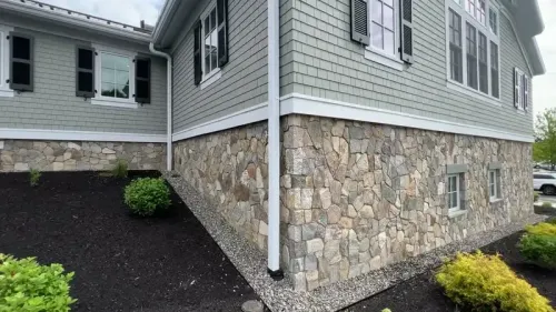House exterior with stone foundation and gray siding, showing a corner and landscaped mulch beds