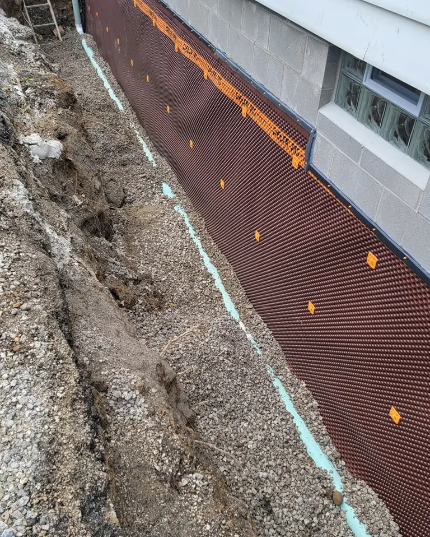 Excavation beside house wall with brown waterproofing membrane and orange drainage markers.