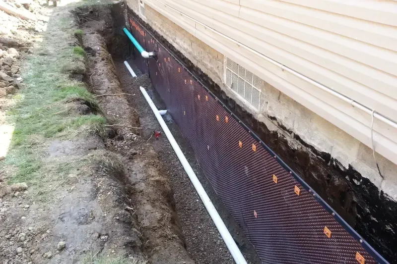 Trench along house foundation with black drainage mat, white pipe, and exposed soil beside beige siding