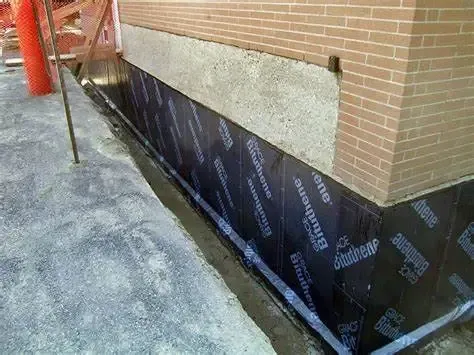 Black waterproofing membrane along a brick wall at the base of a building, beside a concrete walkway.