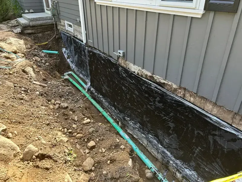 House foundation wrapped in black waterproofing beside a dirt trench and green drainage pipe