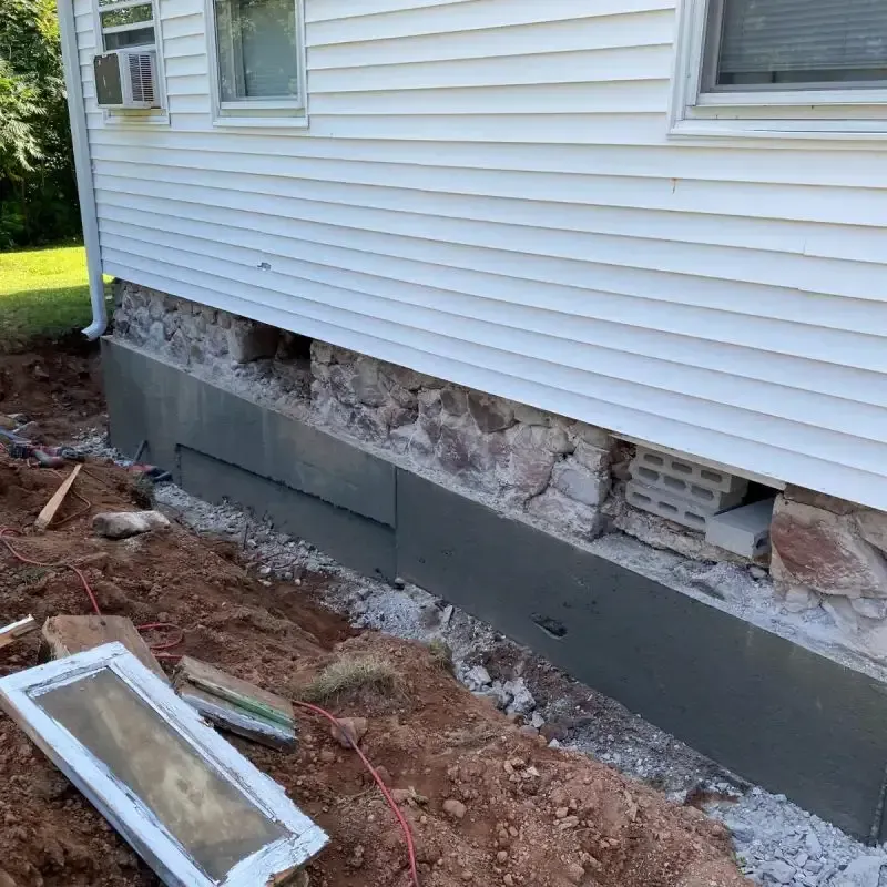 House foundation under repair with exposed stone and gray waterproofing along the base exterior wall.
