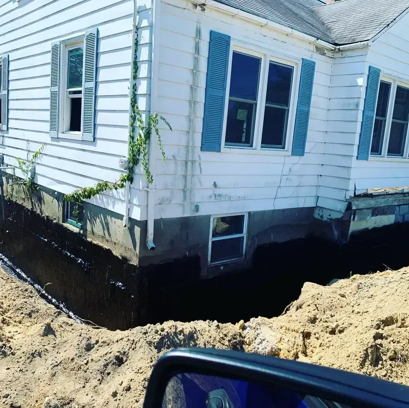 House under construction with white siding and blue shutters, surrounded by excavated dirt.