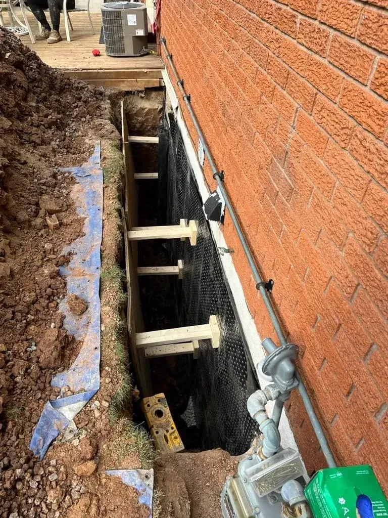Trench beside brick wall with wooden supports, exposed foundation, dirt piles, and pipe running along the wall