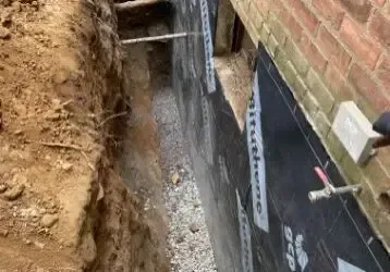 Narrow trench beside a brick house with exposed foundation, waterproofing membrane, and wet soil.