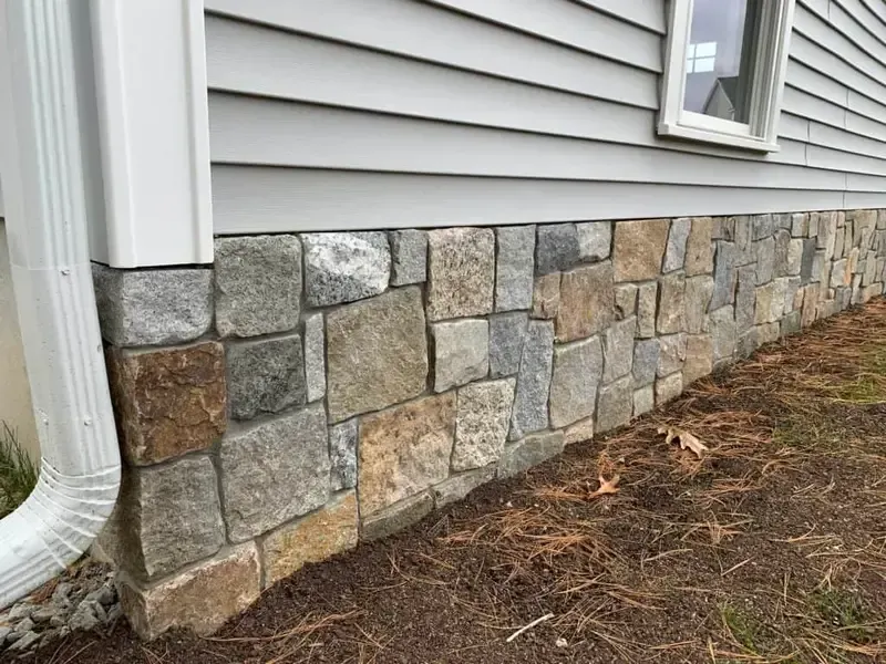 Stone veneer skirt on a house exterior beneath gray siding and a white downspout
