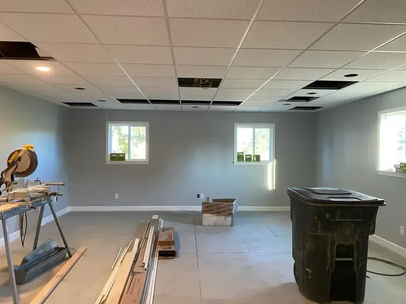 Empty room under renovation with tools, floor boards, trash bin, and ceiling tiles missing
