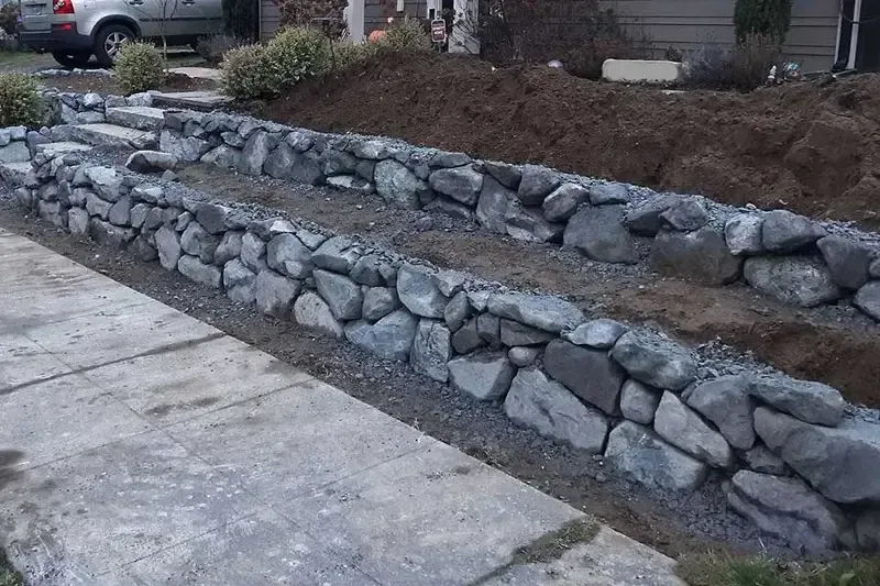 Stone retaining wall beside a concrete driveway with a landscaped yard and parked car in the background