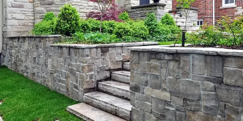 Stone steps built into a raised gray retaining wall beside a green lawn and brick house.
