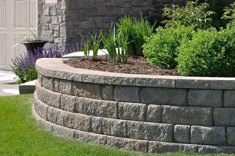 Curved stone retaining wall with mulch and green plants beside a manicured lawn and stone house