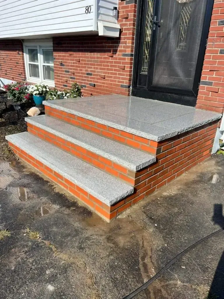 Front steps with gray tile treads and orange trim leading to a black front door on a brick house