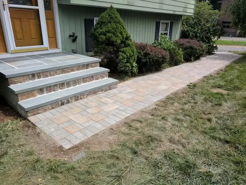 Front steps and a paved walkway leading to a house entrance, with shrubs and a lawn nearby.