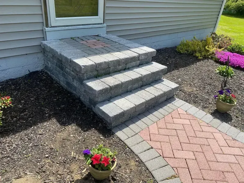 Front porch steps with gray pavers leading to a door, bordered by mulch and small flower pots.
