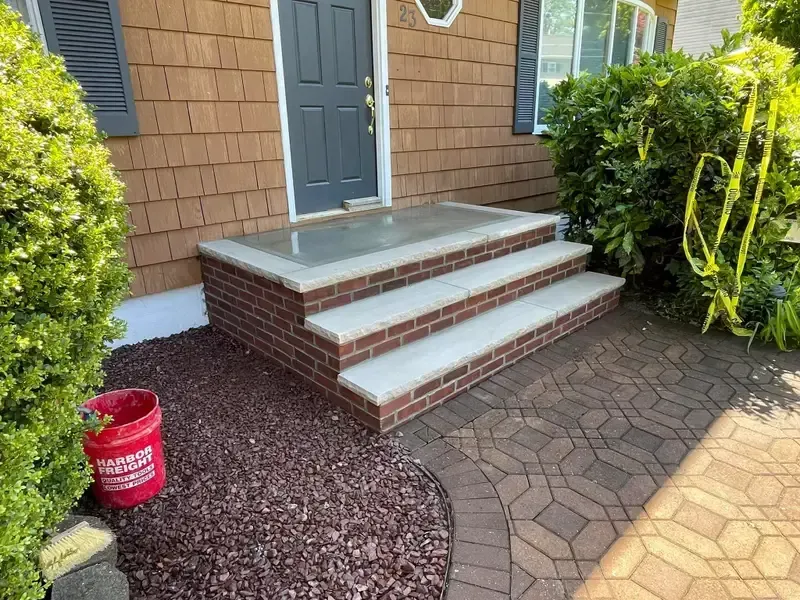 Front porch with three brick steps, gray door, shrubs, and a red bucket on gravel walkway.