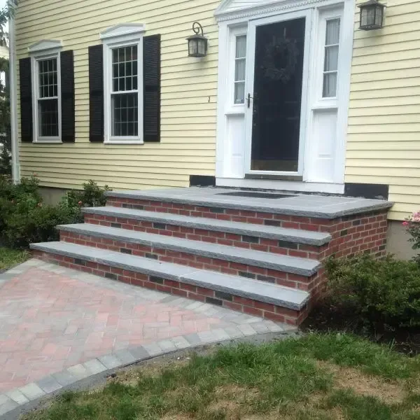 Front steps and white-trimmed black door on a yellow house with brick walkway and porch railing