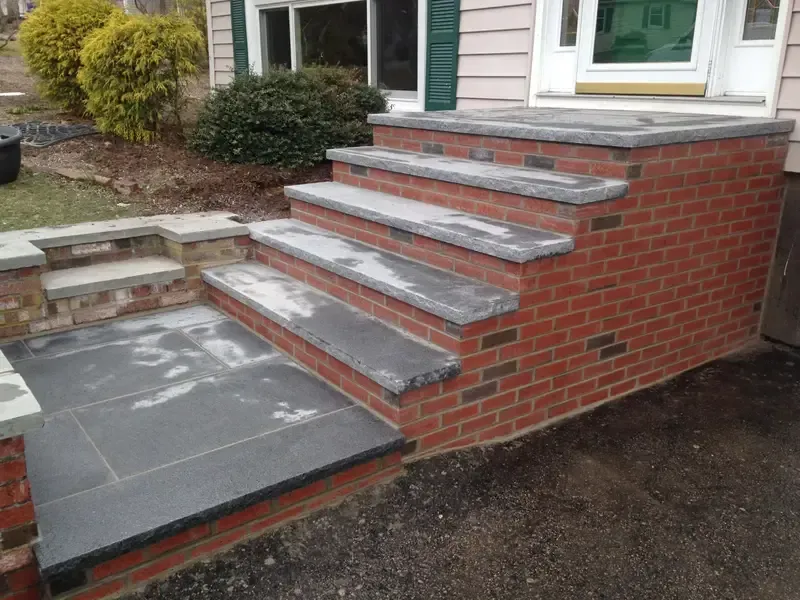 Brick front steps with stone treads leading to a house entrance, beside a small patio.