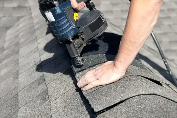 Worker using a roofing nail gun to install gray shingles on a roof