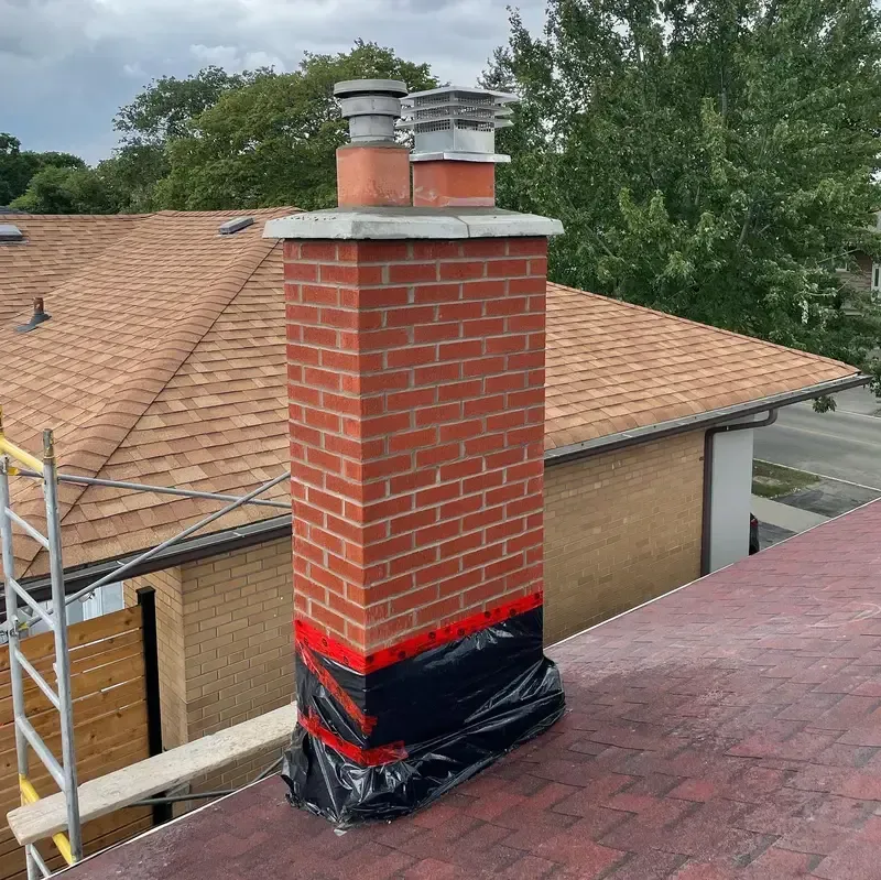 Brick chimney on a rooftop, wrapped at the base with black plastic and red tape.