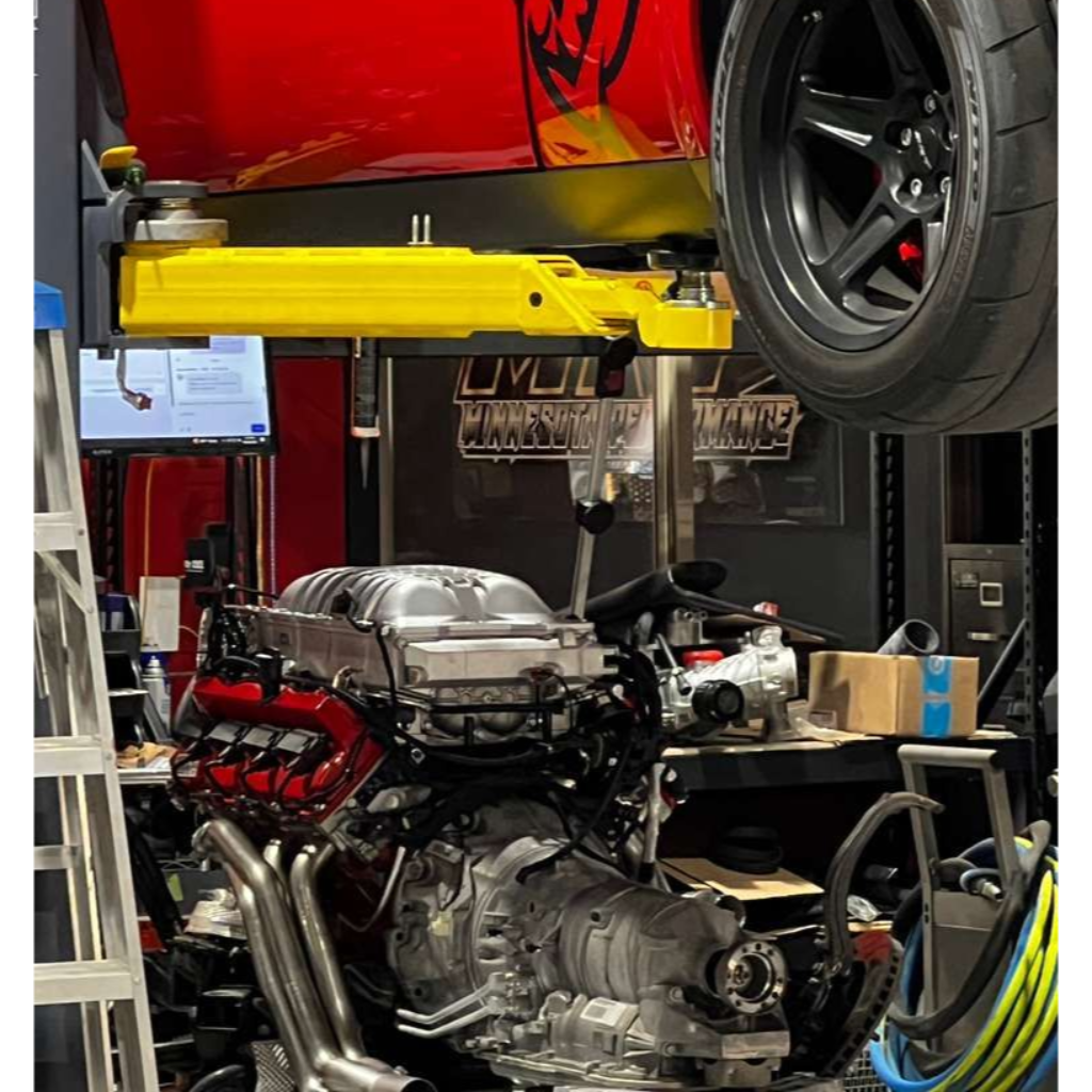 Car battery in an engine bay with red positive terminal, black hold-down bracket, and various components.