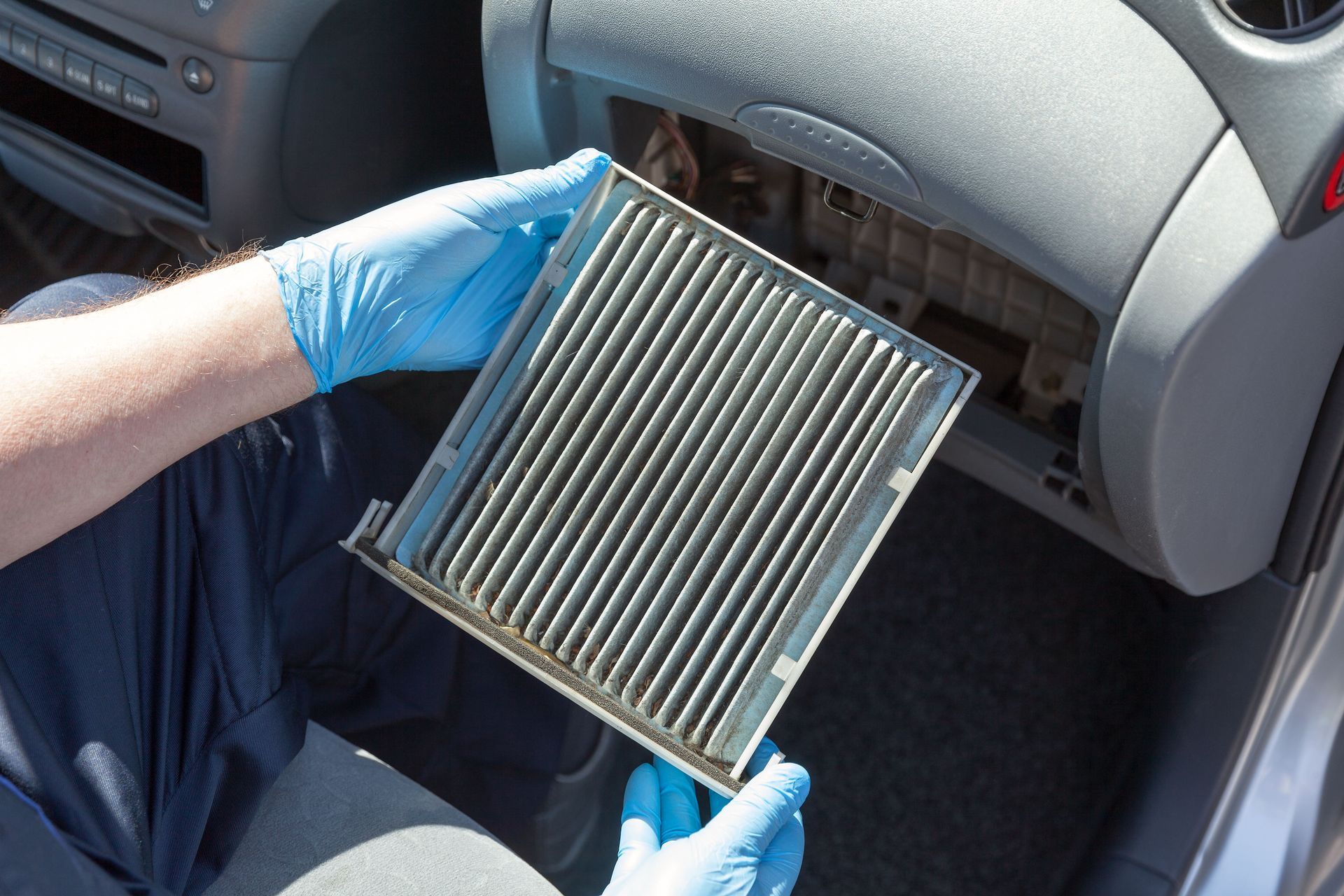 Person in blue gloves holding a dirty, rectangular cabin air filter from a car's dashboard.