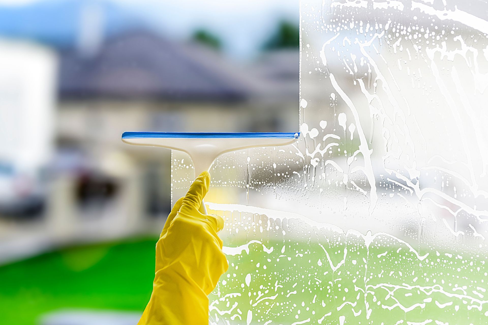 Yellow-gloved hand wiping a window with a blue squeegee, clearing white foam over a blurred house background