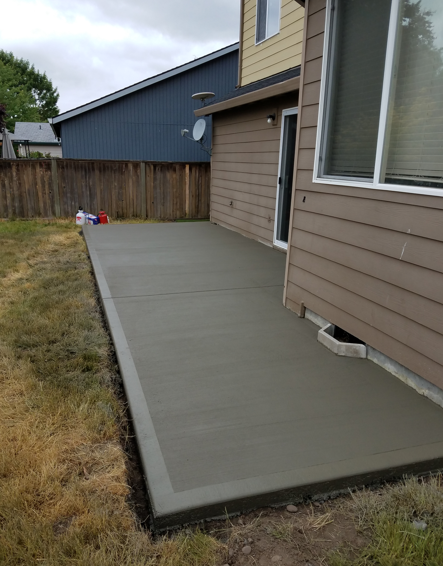Concrete patio beside a house, bordered by grass and a wooden fence, with a sliding glass door.