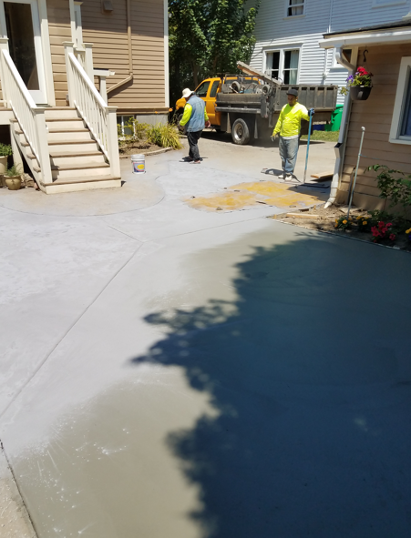 Workers paving a driveway beside homes, with a truck and fresh concrete in a sunny residential area