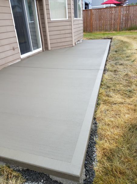 Fresh concrete patio beside a house, bordered by a yard and wooden fence