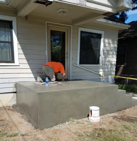 Worker smoothing a freshly poured concrete porch beside a house.