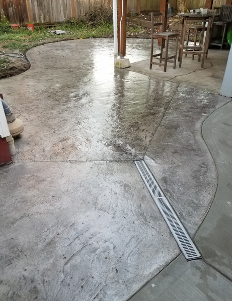 Concrete patio with a narrow drainage grate, wooden fence, and small table and chairs.
