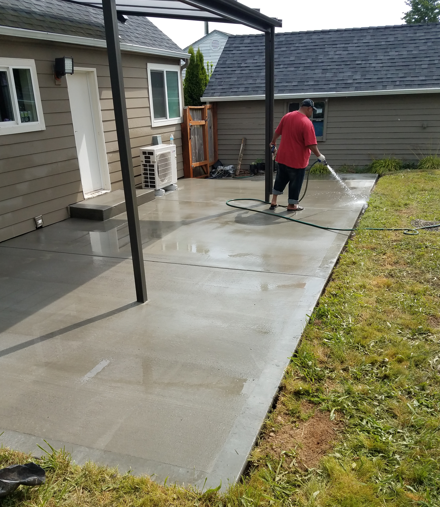 Person pressure-washing a wet concrete patio beside a house and lawn.