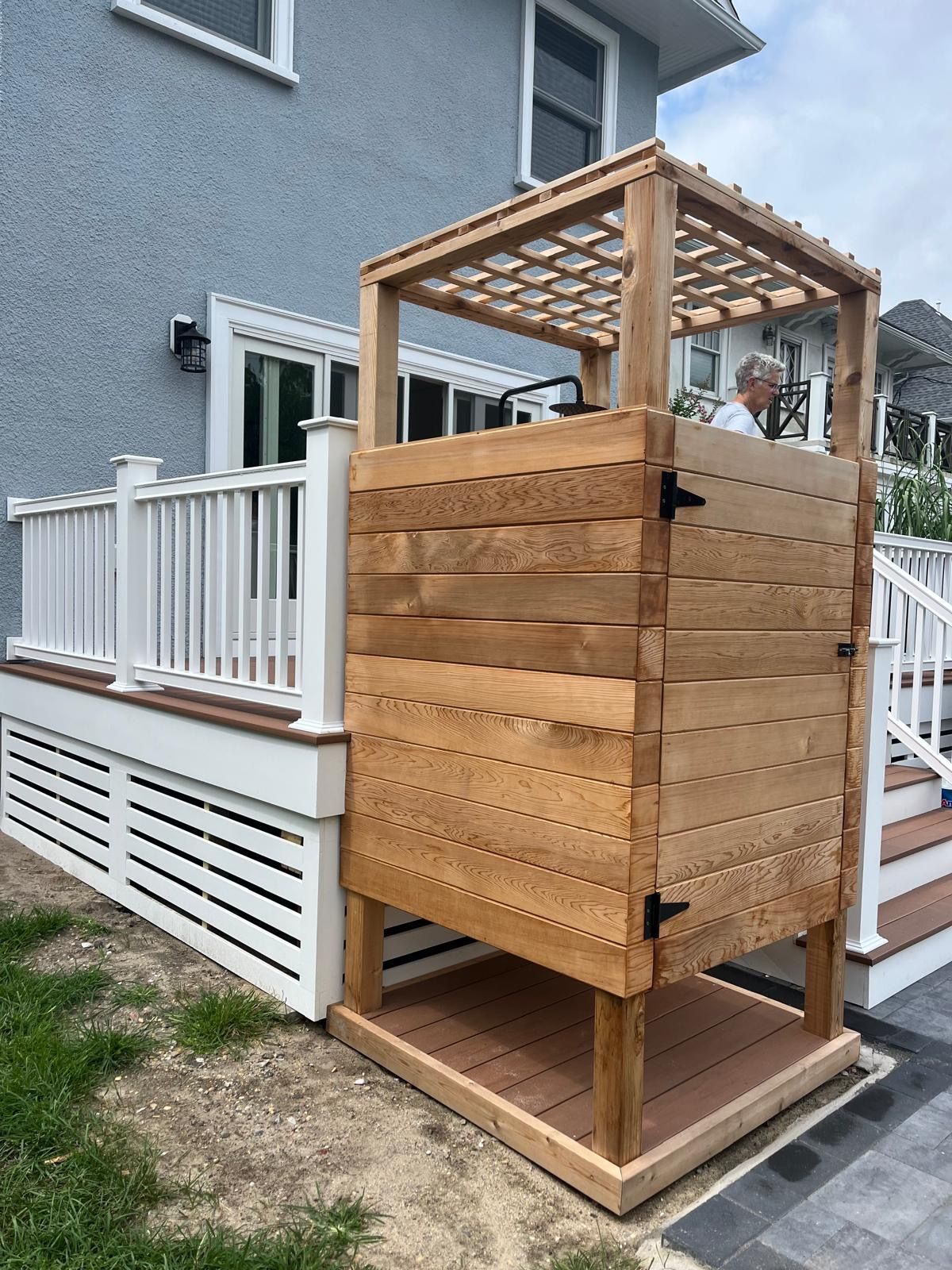 A wooden outdoor shower is sitting on the side of a deck next to a house.