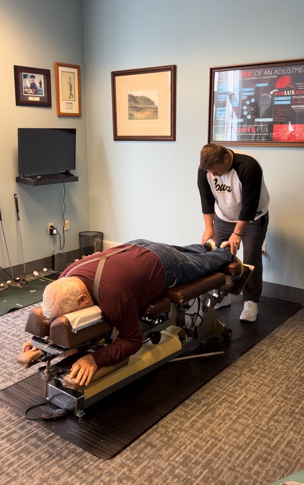 Man lying on chiropractic table as another man adjusts his legs in a room with framed art.