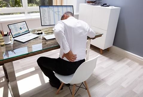 Man seated at desk, touching his lower back in pain. Office setting with laptop and computer monitor.