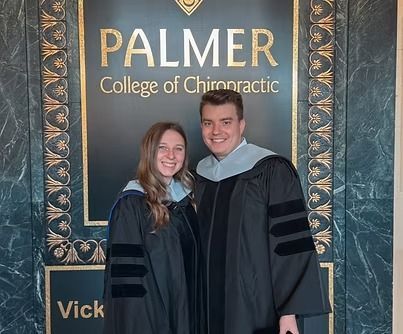 Two people in graduation gowns pose in front of a Palmer College of Chiropractic sign.